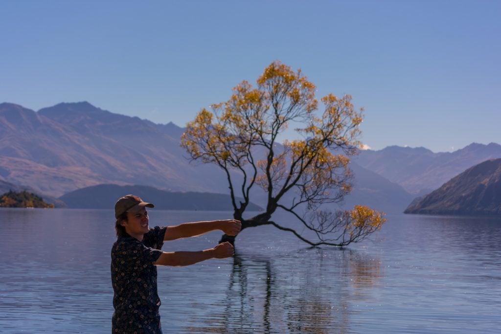 Ryan Schrader stands near the lake in Wanaka New Zealand. Behind him a leafy tree grows right up out of the lake.