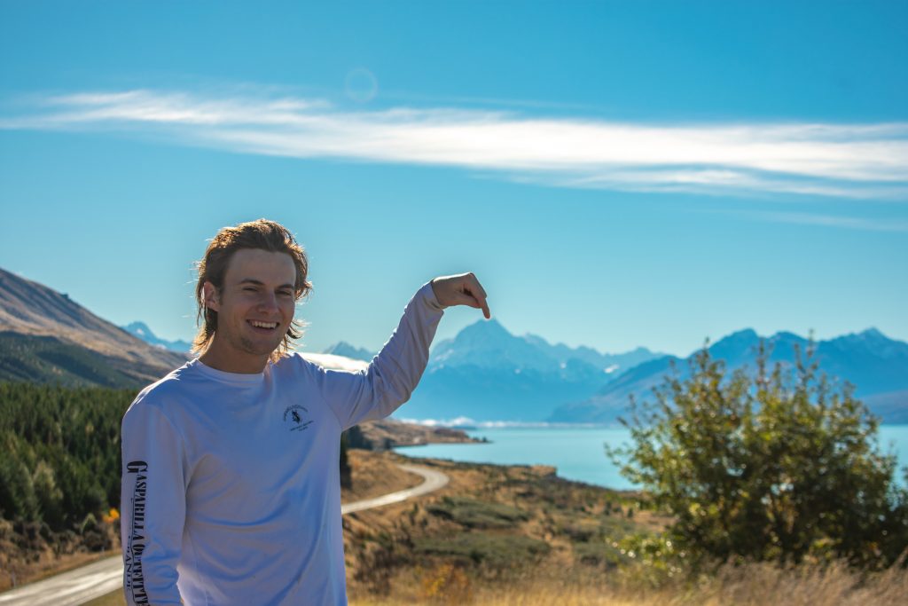 Ryan Schrader at the side of a winding mountain road, pointing at the mountain range in the background. His hand is curved and appears to touch the tip of the mountain.