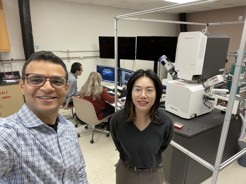 Two people standing in a lab with two others seated at a computer in the background.