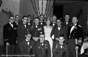 A group dressed in military attire pose for a photo. A woman wearing a corsage stands in the middle of the group.