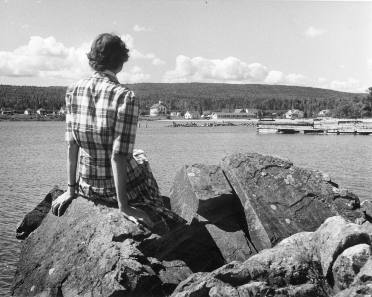 A visitor sitting on a rock enjoys a peaceful reflection at Eagle Harbor Beach in the mid-twentieth century.