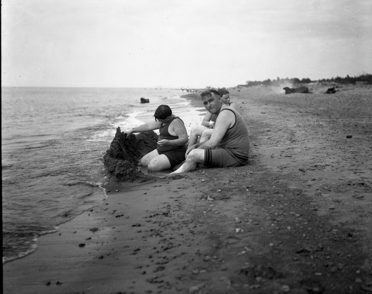 Two people are sitting on a sandy Copper Country beach near the water, with one building a sandcastle.