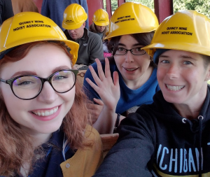 Three women are pictured in a tram car with hard hats on, preparing to descend into the mine tour.