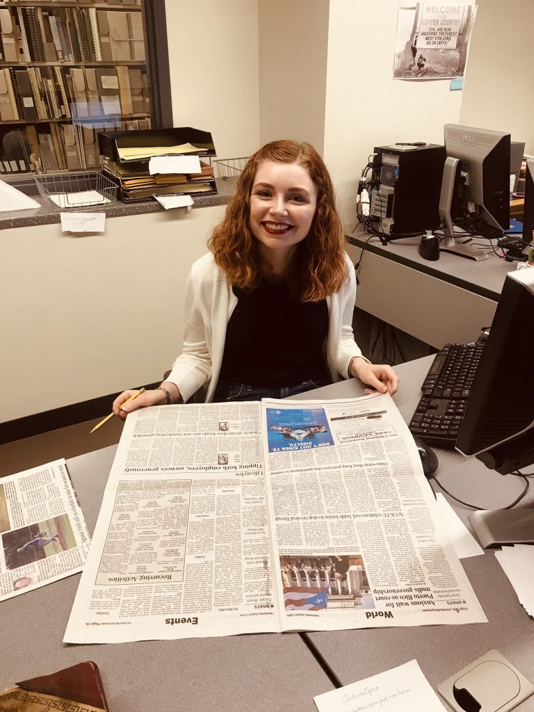 A young woman sits at a desk with a newspaper.