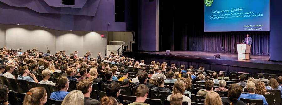 Ronald Jackson stands in front of a podium and large presentation screen on stage while a large audience is seated in front of him.