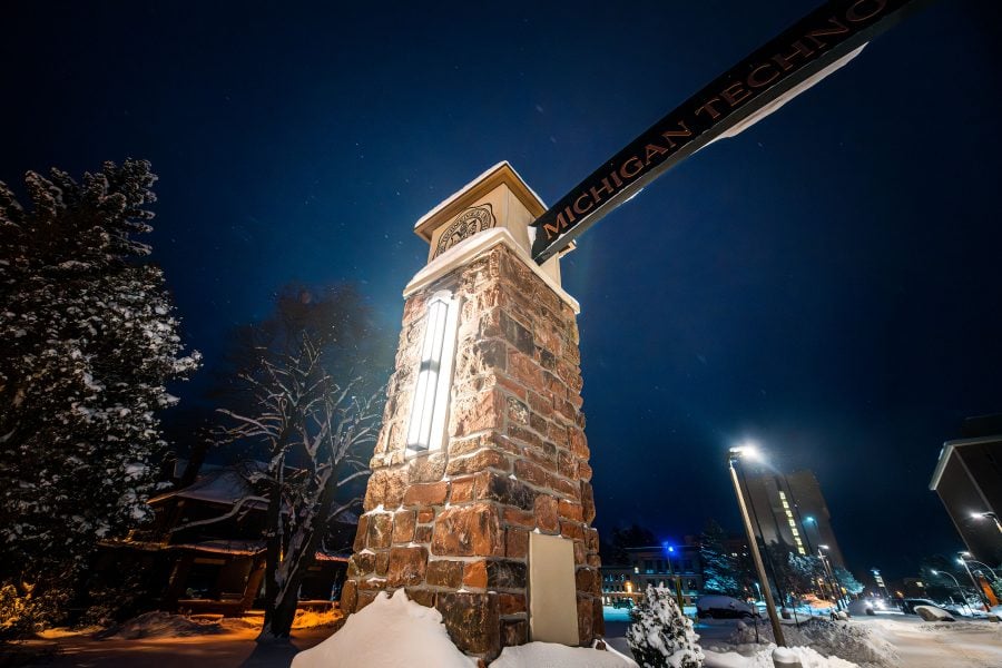 The Michigan Tech Alumni Gateway Arch is lit on a cold winter night with a snowy campus in the background.