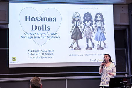 A student stands in front of a screen in a university lecture hall that shows Hosanna Dolls, her project that she is pitching at the Husky Innovate competition at Michigan Tech.