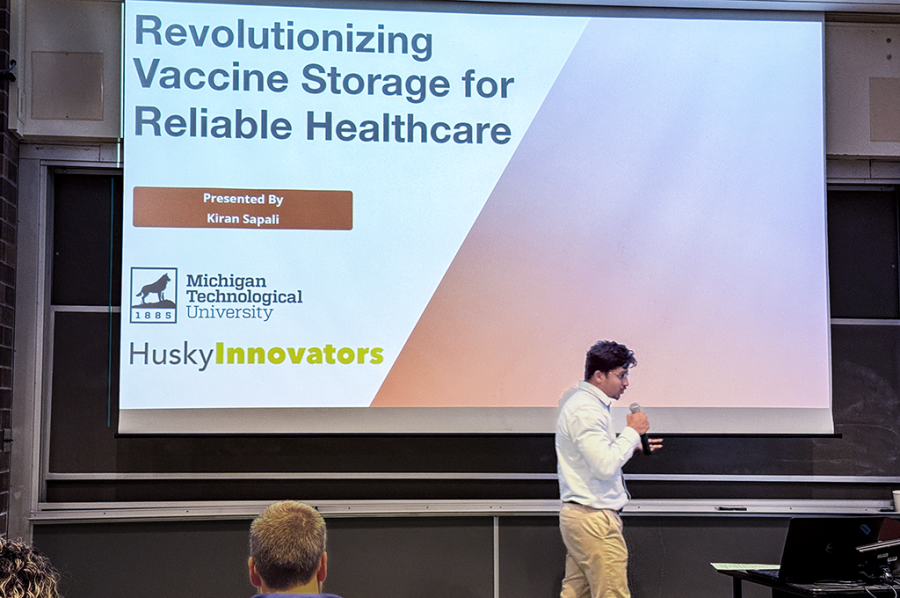 A student with a microphone presents his idea “Revolutionizing Vaccine Storage for Reliable Healthcare” projected on the screen behind him in a lecture hall at Michigan Technological University.