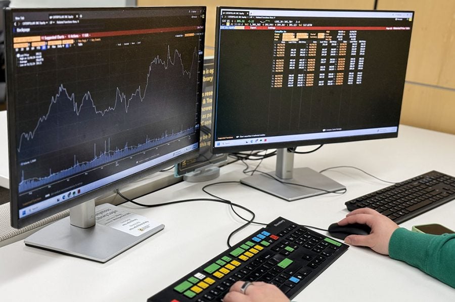 A graph of stock market data with a waveform on a computer screen, stock listings on a second computer screen, and two hands at mouse and keyboard are visible as a Michigan Tech finance student logs in on a Bloomberg Terminal in the campus library.