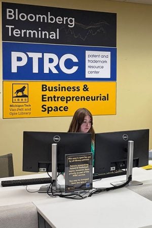 A Michigan Tech student sits behind a computer at a Bloomberg Terminal with signs behind her that point out the terminal, patent and trademark resource center, and the Michigan Tech Business and Entrepreneurial Space. There is a desk with seating in front of the terminal.