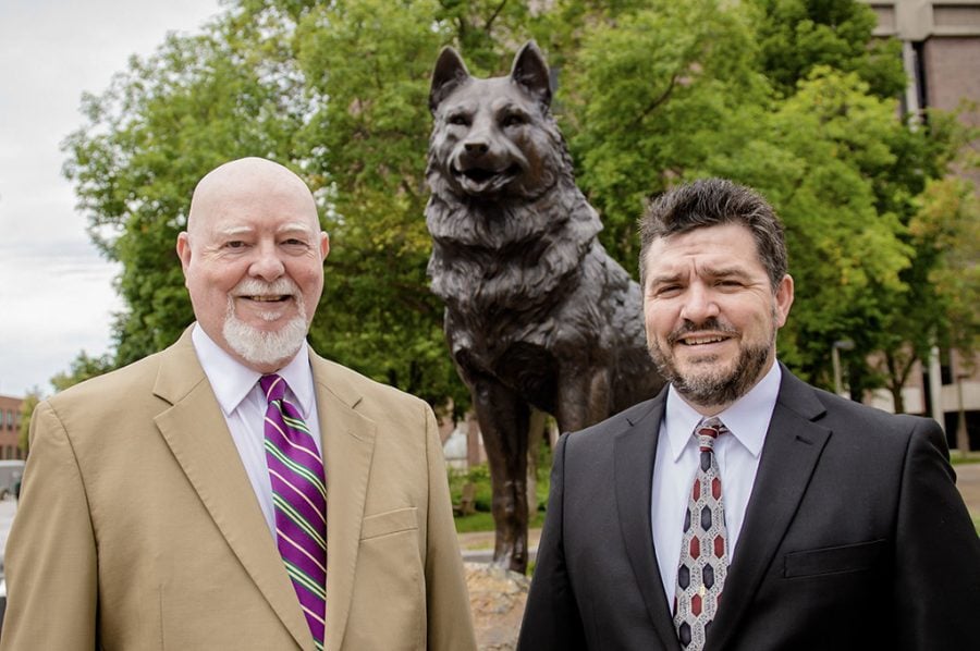 Two Michigan Tech Deans in suits shake hands and smile in front of the Michigan Tech Husky statue with campus buildings in the background in 2015.