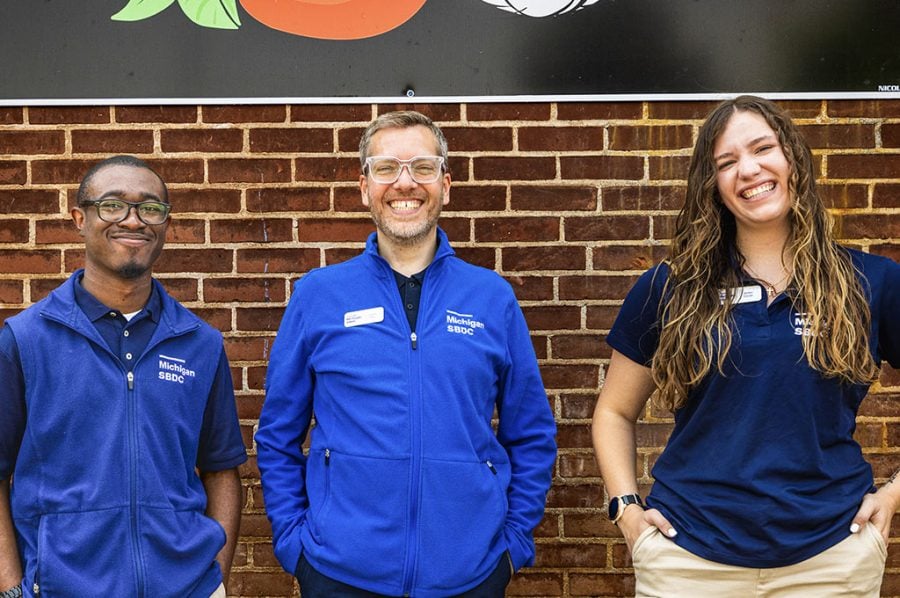 Three people with SBDC shirts are all smiles as they stand in front of a brick wall bearing a partially visible logo for Dina Mia Kitchens.