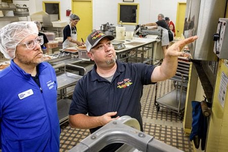 A Michigan SBDC regional associate director looks on as a business owner from Dina Mia Kitchens with logo on his hat and shirt shows a timer on kitchen equipment, with three employees working on sauced pizzas and dough in the background.