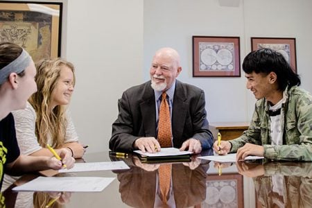 A dean in the Michigan Tech College of Business sits with three students in a conference room at a reflective table as they smile and talk with folders, papers, and pens.