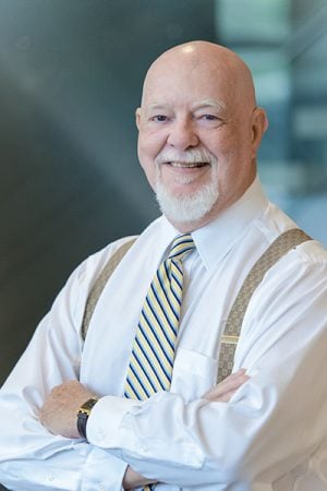 A man in suspenders with a neutral blue background smiles at the camera.