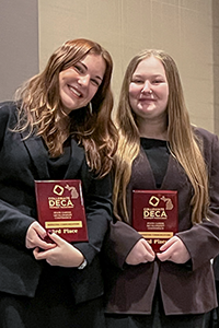 Two smiling young women who are Michigan Tech students hold DECA award plaques as a row of other students stand behind them near a table of awards at the Michigan Collegiate conference competition.