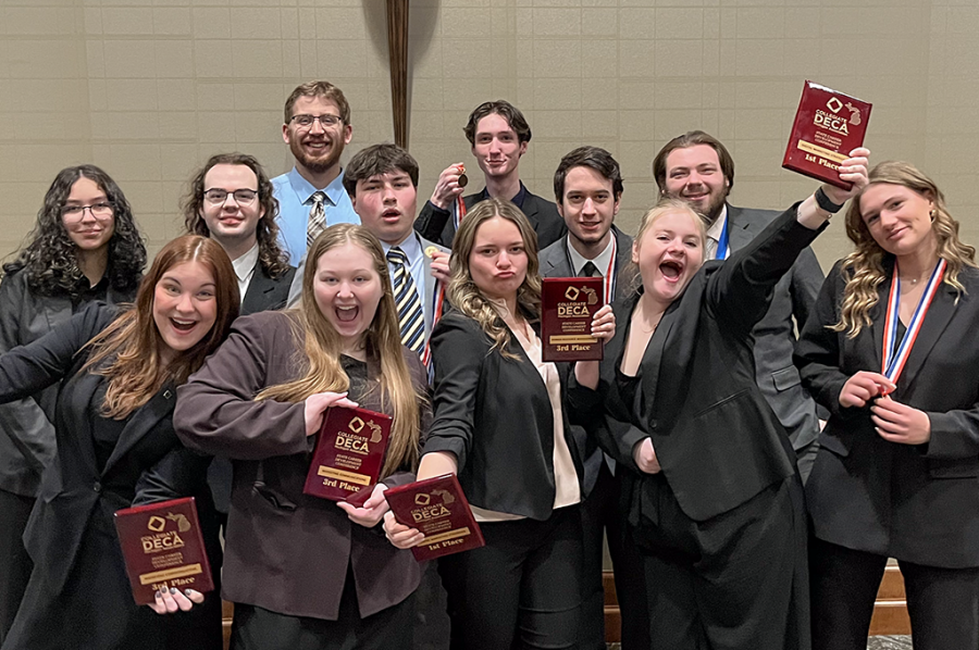 12 Michigan Tech students in suits joke around and hold up DECA awards in front of a beige wall at a statewide conference.