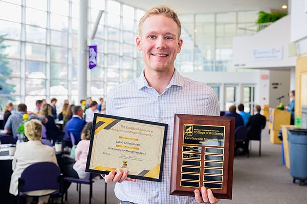 A student holds a plaque and a framed certificate for a construction management award at the College of Business awards ceremony at Michigan Tech. Students, faculty, and alumni are in the background in a banquet setting in the lobby of the Rozsa Center.