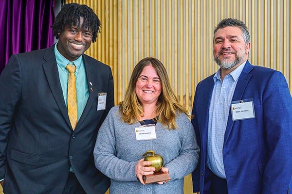 A student, teacher, and college dean stand in front of a wood wall with a purple-curtained backdrop during an awards ceremony for the Michigan Tech College of Business. The teacher is holding a golden apple as winner of the teacher of the year. Their name tags read Elijah Joseph, Laura Sieders, and Dean Johnson.