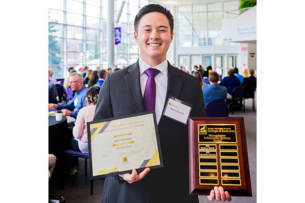 A student smiling in suit and tie holds a wooden plaque and a framed award certification for the 2025 College of Business Engineering Management Award at the annual awards ceremony.