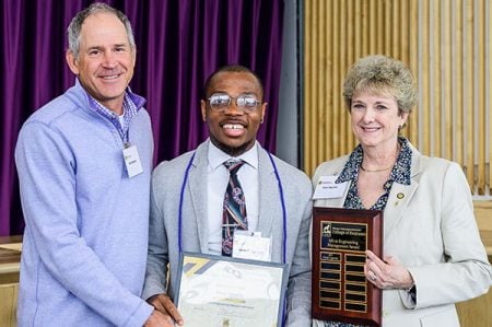 A student holds a plaque and smiles as they receive a Michigan Tech College of Business Award and a distinguished alumni and member of college leadership look on.
