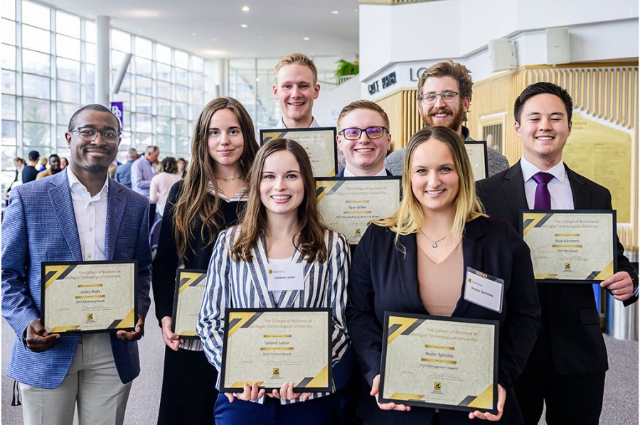 Eight students stand smiling with framed awards certificates at the annual College of Business ceremony and banquet in the Rozsa Center at Michigan Tech. Behind them, faculty, staff, alumni, and student attendees stand and sit at tables in the lobby, mingling.