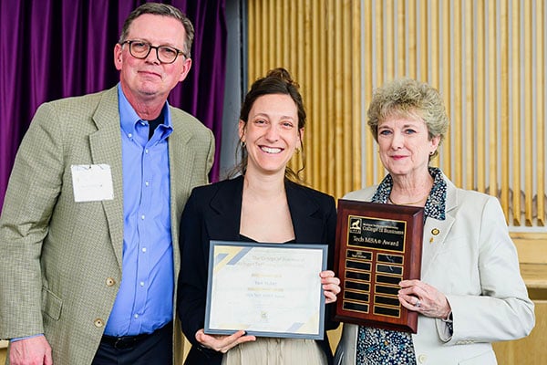 A student holds a plaque A student holds a plaque and smiles as they receive a Michigan Tech College of Business Award and a distinguished alumni and member of college leadership look on.