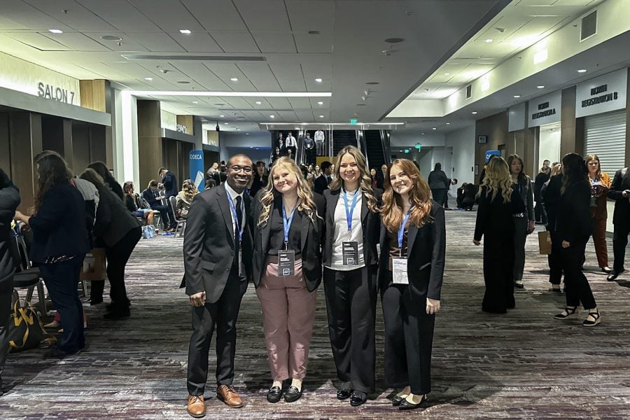 Four Michigan Tech DECA students attending the Collegiate DECA ICDC 2025 event in San Francisco stand in the center of a reception hall with other students, also wearing lanyards and business attire, registering and networking around them.