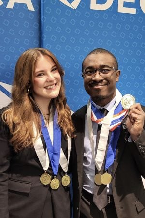 Two smiling students wearing medals on ribbons around their necks and professional business attire stand in front of a DECA sign at a conference where they competed.