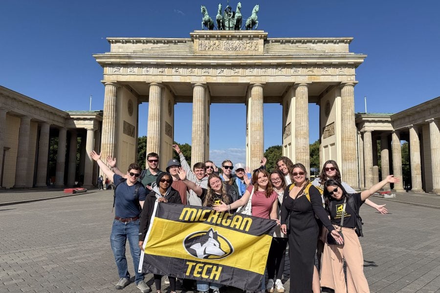 A group of Michigan Tech students hold a Husky flag in front of a historic building in Germany during a study trip focused on supply chain and lean manufacturing.