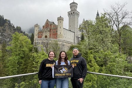 Three Michigan Tech students wearing Michigan Tech shirts and holding a Michigan Tech Huskies flag stand in front of Neuschwanstein Castle