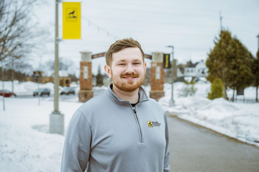 A student wearing a Michigan Technological University shirt stands under the Alumni Gateway Arch at Michigan Tech as he prepares to collect his second degree from the institution.