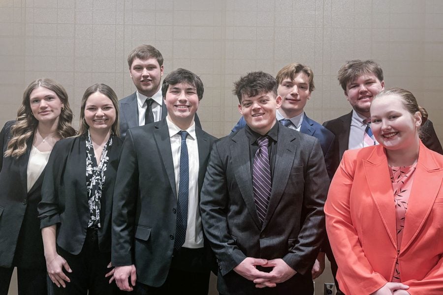 Eight smiling Michigan Tech College of Business students in business attire pose for a photo together with a blank wall behind them at the state DECA conference in lower Michigan.