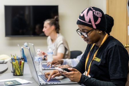 Two Michigan Tech students, one with a Husky EATS logo on her shirt wearing a black and pink hat and a lanyard, stare intently at the screens of phones and laptops as they work on taxes for community members. The suit and lanyard of their faculty supervisor is visible on the right.