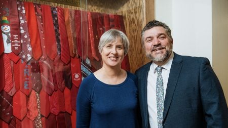 Two professors in the Michigan Tech College of Business smile in front of a glass case displaying red ties sent to the College by their business students who have earned their CPA credentials.