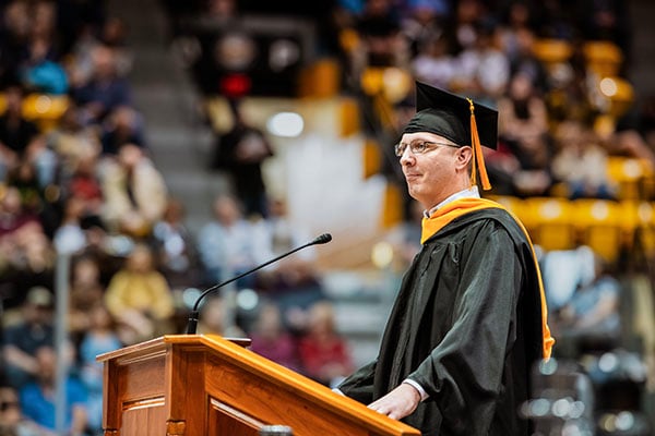 A smiling Michigan Tech alumnus in cap and gown stands at a podium at graduation in the MacInnes Ice Arena addressing new graduates