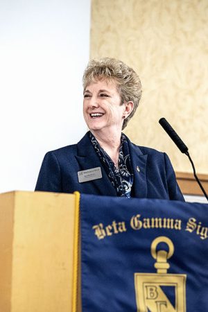 A smiling professor stands at a ballroom podium interacting with students and fellow faculty members at an awards ceremony for the Michigan Tech College of Business.
