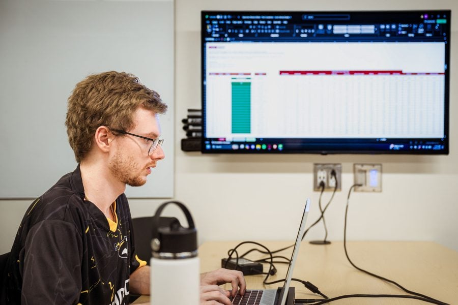 A male student works at a laptop with a wall monitor in the background displaying a spreadsheet.