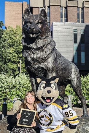 A Michigan Tech Husky poses with a 1st Day of School 2025 sign in front of the University’s iconic bronze Husky statue with the school mascot, Blizzard T. Husky, with their arm around her.