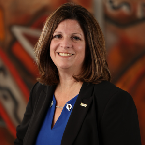 Kimberly Zimmer-Janeczko smiles for a professional portrait wearing a black blazer and royal blue blouse, with Michigan Tech and MDOT pins on her lapel, photographed against a warm-toned abstract background during the 2025 CEGE Academy Induction Ceremony.