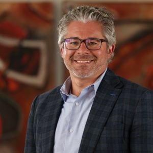 Matthew Bissett smiles for a professional portrait wearing a blue checkered blazer, light blue shirt, and glasses, photographed against a warm-toned abstract background during the 2025 CEGE Academy Induction Ceremony at Michigan Tech.