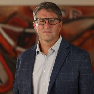 Michael Erickson poses for a professional portrait wearing a gray collared shirt and blue plaid jacket, standing before a warm-toned abstract background during the 2025 CEGE Academy Induction Ceremony at Michigan Tech.
