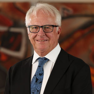 Mike Horan smiles for a professional portrait wearing a black suit jacket, white shirt, and blue patterned tie, with a warm-toned abstract background behind him during the 2025 CEGE Academy Induction Ceremony at Michigan Tech.