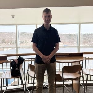 Ryan Keen stands smiling in a bright indoor space with large windows overlooking a snowy waterfront landscape. He is wearing a dark polo shirt, khaki pants, and black shoes, with his hands clasped in front of him. Chairs and high-top tables are visible around him.