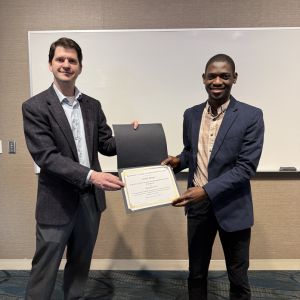 Andrew Swartz presents an award certificate to Israel Adeoye during the CEGE Departmental Awards Ceremony. Israel, wearing a navy blazer, light shirt, dark pants, and sneakers, smiles while holding the certificate. Andrew stands beside him in a blazer and dress shirt in front of a whiteboard in a classroom setting.