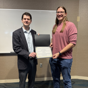 Andrew Swartz presents an award certificate to Kris Crawford during the CEGE Departmental Awards Ceremony. Kris, wearing a pink long-sleeve shirt and jeans, smiles while holding the certificate. Andrew stands beside him in a blazer and dress shirt in front of a whiteboard in a classroom setting.