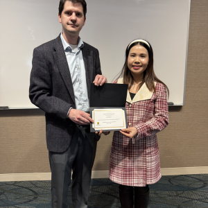 Andrew Swartz presents an award certificate to Linh Nguyen during the CEGE Departmental Awards Ceremony. Linh, wearing a red plaid coat, black boots, and a white headband, smiles while holding the certificate. Andrew stands beside her in a blazer and dress shirt in front of a whiteboard in a classroom setting.