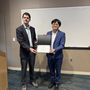 Andrew Swartz presents an award certificate to Meng Wu during the CEGE Departmental Awards Ceremony. Meng, wearing a blue suit and glasses, smiles while holding the certificate. Andrew stands beside him in a blazer and dress shirt in front of a whiteboard in a classroom setting.