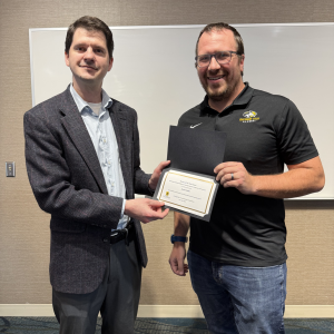 Andrew Swartz presents an award certificate to Zack Fredin during the CEGE Departmental Awards Ceremony. Zack, wearing a black Michigan Tech polo shirt and jeans, smiles while holding the certificate. Andrew stands beside him in a blazer and dress shirt in front of a whiteboard in a classroom setting.