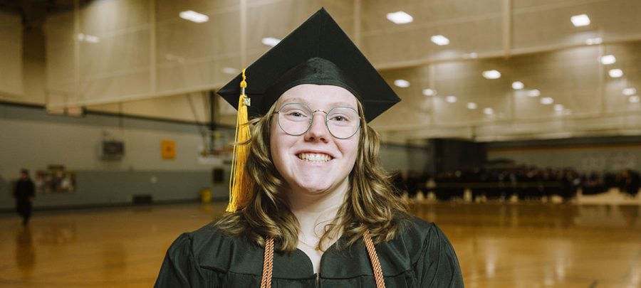 A smiling Michigan Tech graduate in her cap and gown with other graduates gathered behind her as commencement line-up takes place in the university’s multipurpose room.